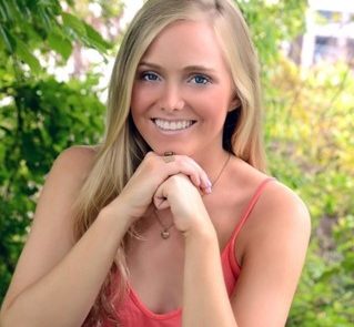 Smiling woman in coral top seated outdoors with greenery in background.