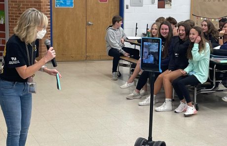 A woman speaks to a seated group with a telepresence robot in front.