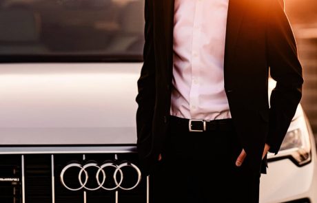 Man in a black suit stands near a white Audi car at sunset.