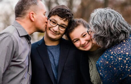 Two adults kissing the cheeks of two smiling children outdoors.