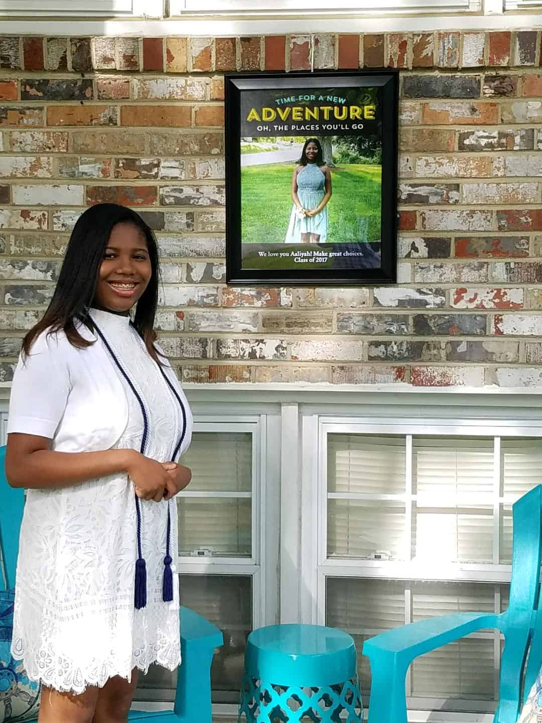 A woman in graduation attire standing in front of a motivational poster.